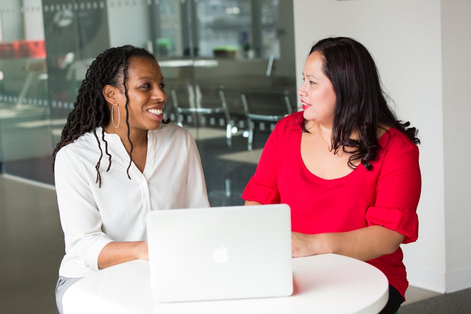Two women in an office discussing work using a laptop, smiling and engaged.